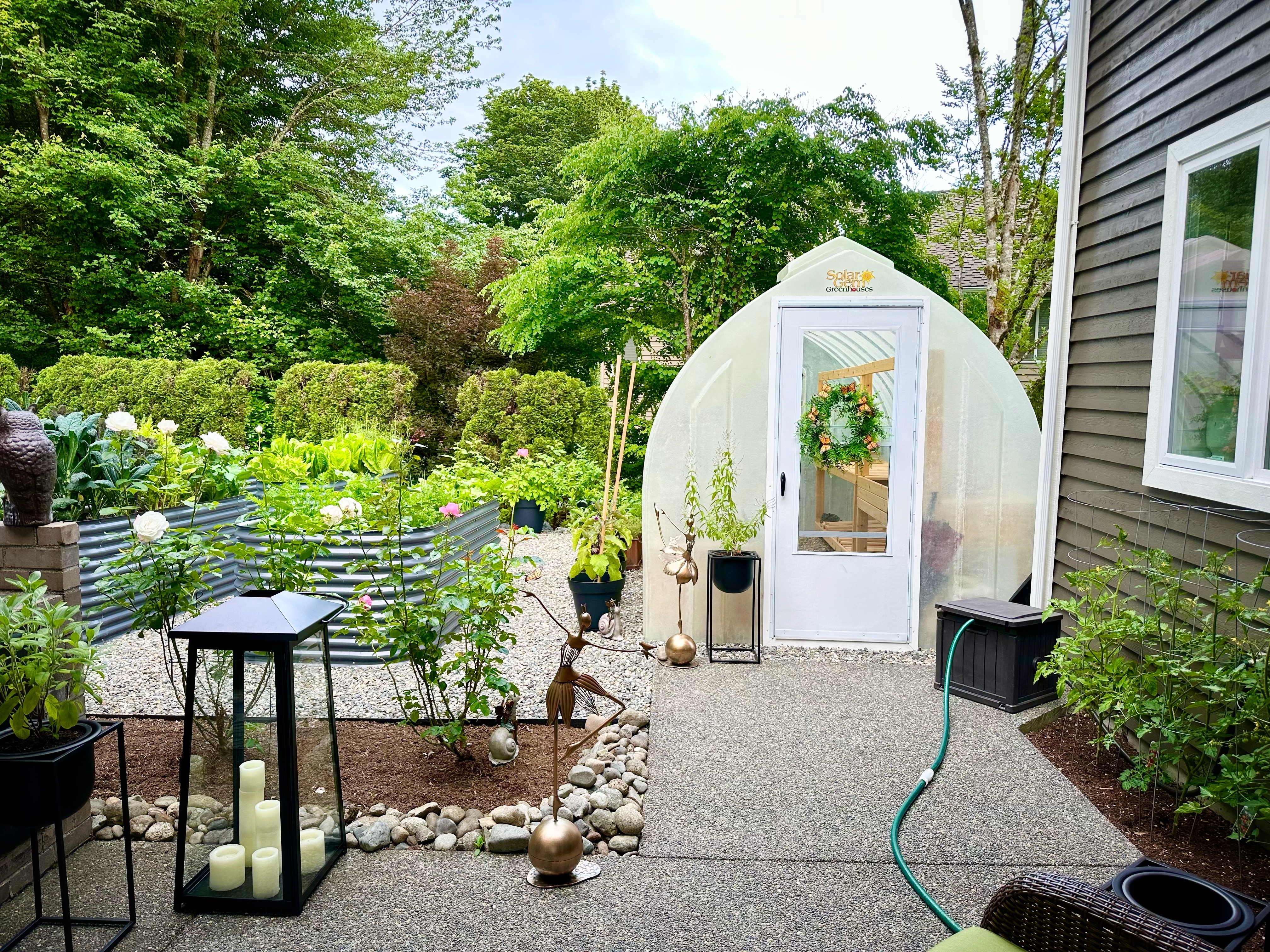 A backyard greenhouse with a wreath on the door, surrounded by raised garden beds, potted plants, and decor on a gravel and concrete patio.